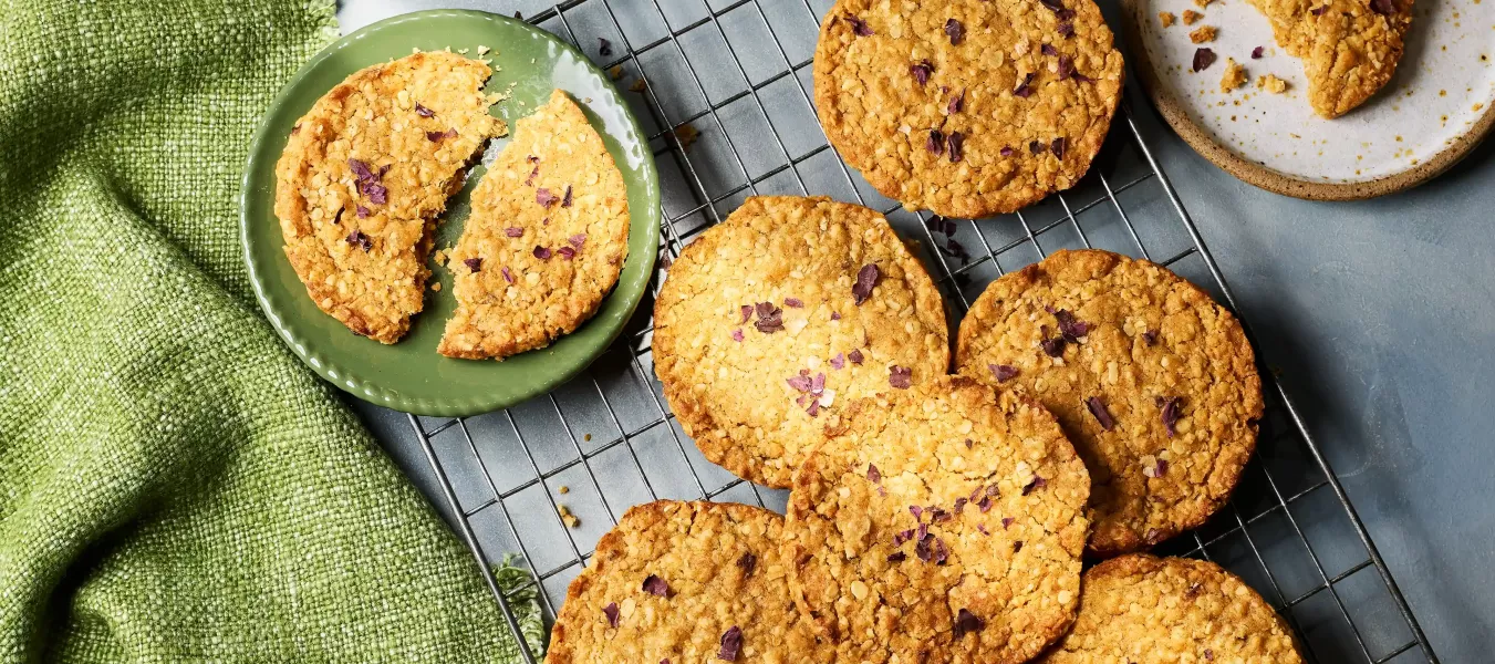 Mehrere runde, goldbraun gebackene Hafercookies liegen auf einem Abkühlgitter. Ein Teller mit einem in zwei Hälften gebrochenen Cookie steht daneben.