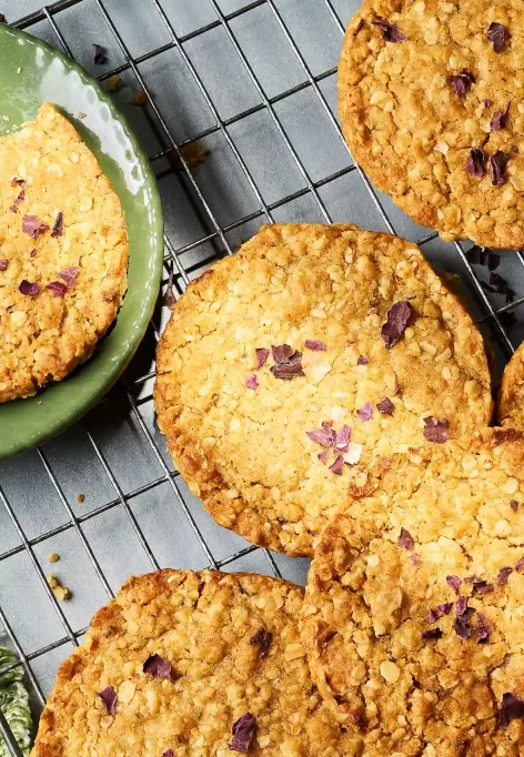 Mehrere runde, goldbraun gebackene Hafercookies liegen auf einem Abkühlgitter. Ein Teller mit einem in zwei Hälften gebrochenen Cookie steht daneben.