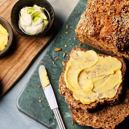 Ein angeschnittenes dunkles Brot liegt auf einem Brett; eine Scheibe ist mit gelber Butter bestrichen. Daneben stehen drei kleine Schalen mit unterschiedlichen Buttersorten sowie ein Messer.