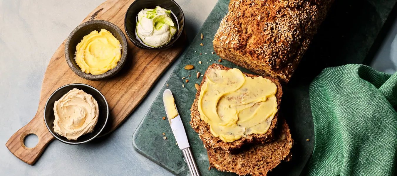 Ein angeschnittenes dunkles Brot liegt auf einem Brett; eine Scheibe ist mit gelber Butter bestrichen. Daneben stehen drei kleine Schalen mit unterschiedlichen Buttersorten sowie ein Messer.