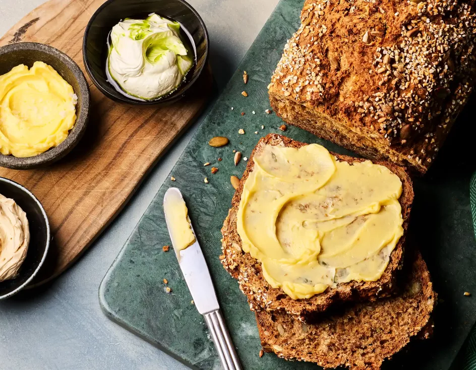 Ein angeschnittenes dunkles Brot liegt auf einem Brett; eine Scheibe ist mit gelber Butter bestrichen. Daneben stehen drei kleine Schalen mit unterschiedlichen Buttersorten sowie ein Messer.