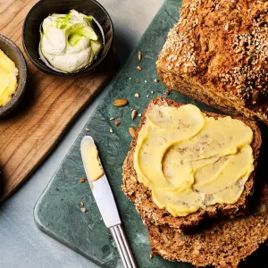 Ein angeschnittenes dunkles Brot liegt auf einem Brett; eine Scheibe ist mit gelber Butter bestrichen. Daneben stehen drei kleine Schalen mit unterschiedlichen Buttersorten sowie ein Messer.