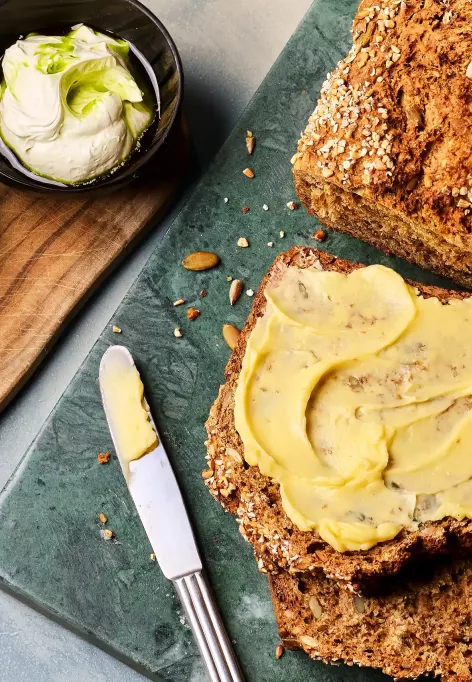 Ein angeschnittenes dunkles Brot liegt auf einem Brett; eine Scheibe ist mit gelber Butter bestrichen. Daneben stehen drei kleine Schalen mit unterschiedlichen Buttersorten sowie ein Messer.