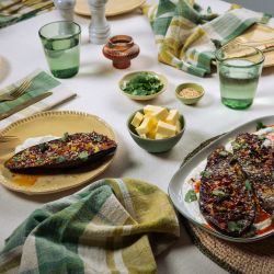 Ein gedeckter Tisch mit gebratenen Auberginen auf Joghurt, umgeben von kleinen Schalen mit Kräutern, Butter und Sesam sowie Gläsern, Tellern und Stoffservietten.
