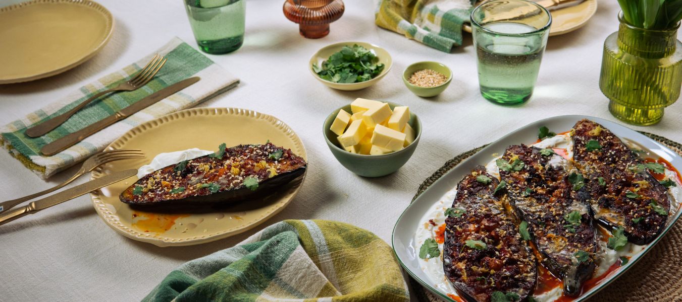 Ein gedeckter Tisch mit gebratenen Auberginen auf Joghurt, umgeben von kleinen Schalen mit Kräutern, Butter und Sesam sowie Gläsern, Tellern und Stoffservietten.