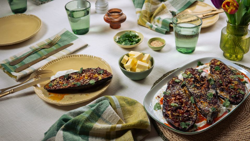 Ein gedeckter Tisch mit gebratenen Auberginen auf Joghurt, umgeben von kleinen Schalen mit Kräutern, Butter und Sesam sowie Gläsern, Tellern und Stoffservietten.