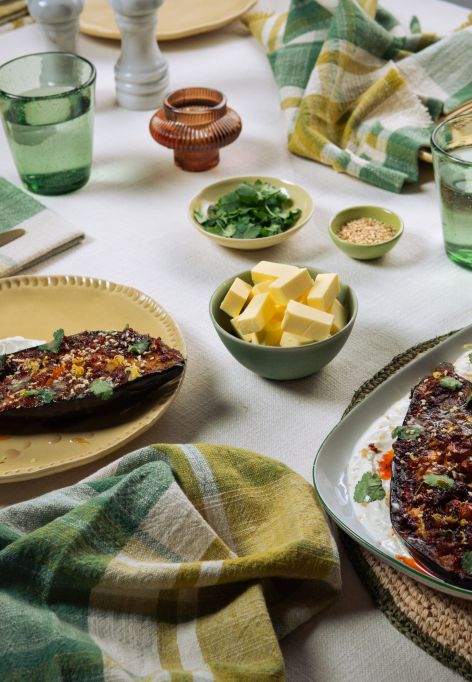 Ein gedeckter Tisch mit gebratenen Auberginen auf Joghurt, umgeben von kleinen Schalen mit Kräutern, Butter und Sesam sowie Gläsern, Tellern und Stoffservietten.