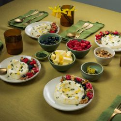 Auf einem gedeckten Tisch stehen mehrere Teller mit weißer Mousse, garniert mit Beeren und Nüssen. Daneben befinden sich kleine Schalen mit Butterstücken, Beeren und Nüssen sowie braune Gläser und grüne Stoffservietten.