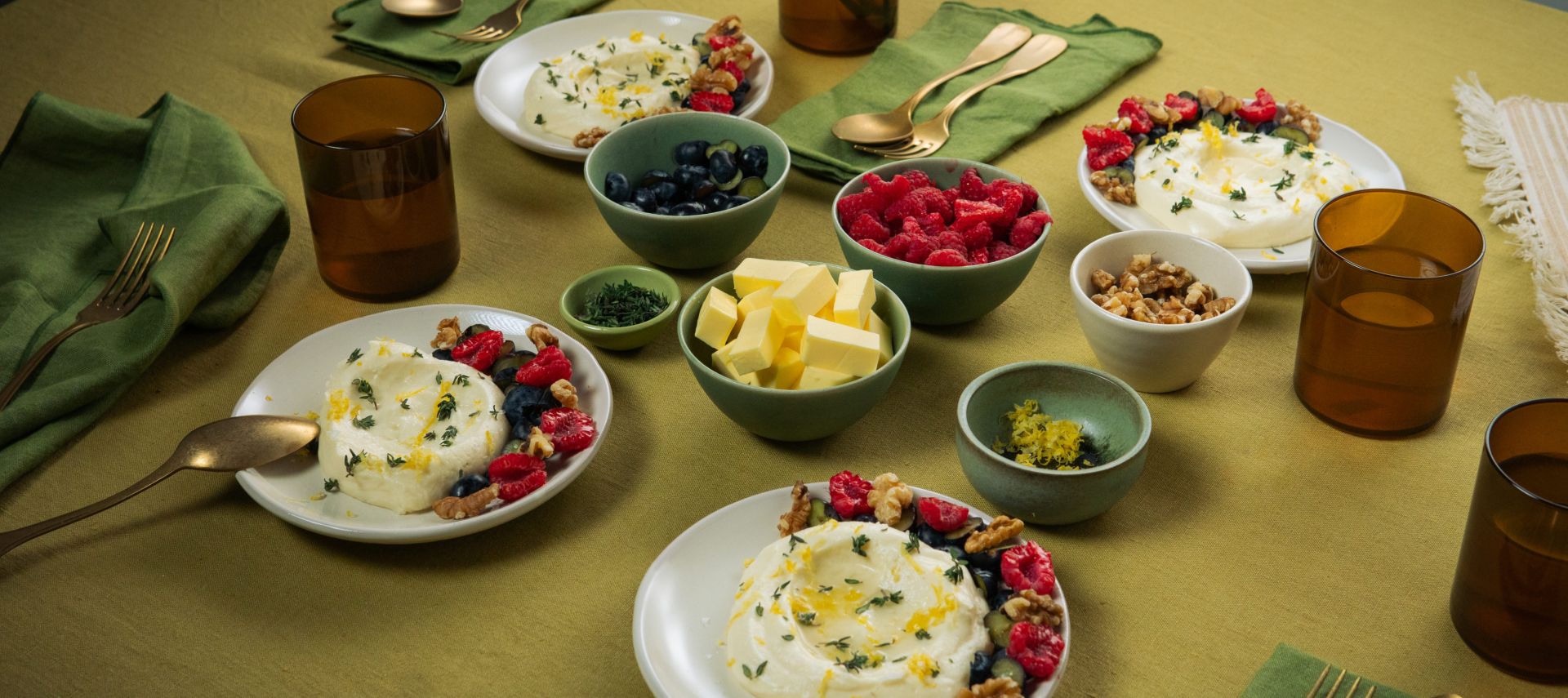 Auf einem gedeckten Tisch stehen mehrere Teller mit weißer Mousse, garniert mit Beeren und Nüssen. Daneben befinden sich kleine Schalen mit Butterstücken, Beeren und Nüssen sowie braune Gläser und grüne Stoffservietten.