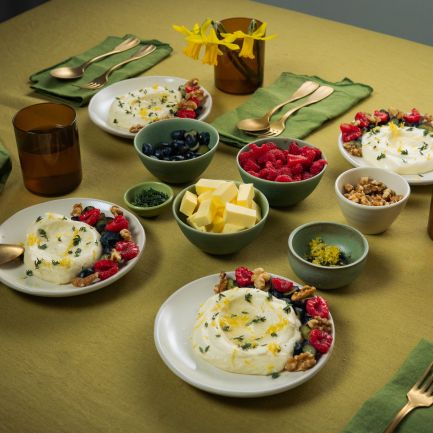 Auf einem gedeckten Tisch stehen mehrere Teller mit weißer Mousse, garniert mit Beeren und Nüssen. Daneben befinden sich kleine Schalen mit Butterstücken, Beeren und Nüssen sowie braune Gläser und grüne Stoffservietten.