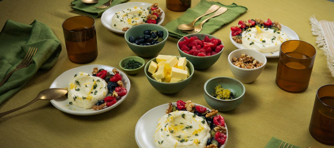 Auf einem gedeckten Tisch stehen mehrere Teller mit weißer Mousse, garniert mit Beeren und Nüssen. Daneben befinden sich kleine Schalen mit Butterstücken, Beeren und Nüssen sowie braune Gläser und grüne Stoffservietten.