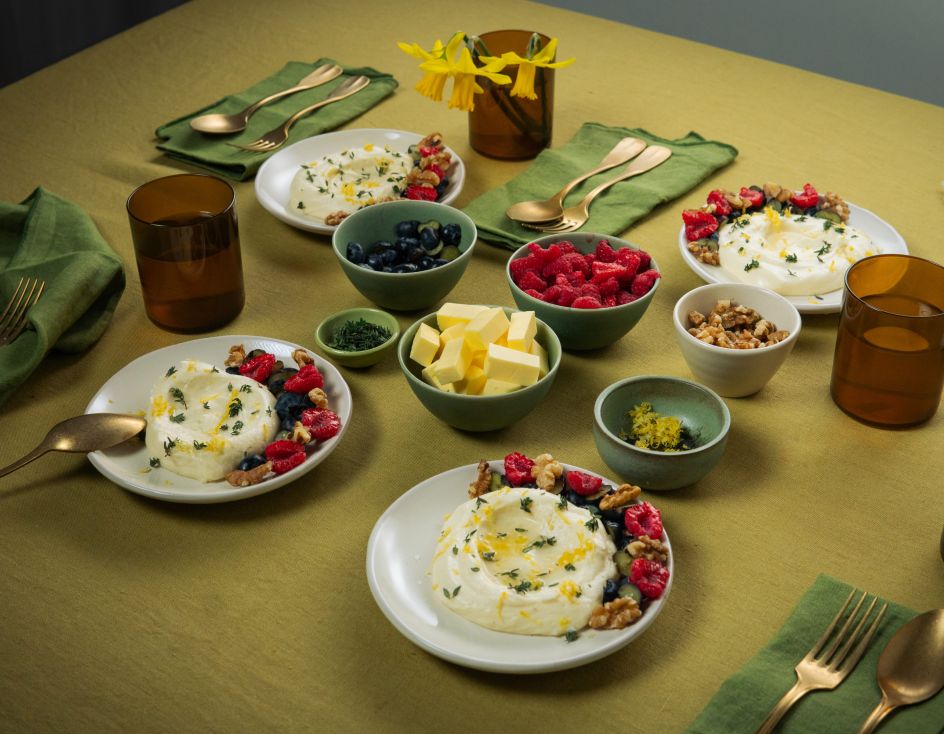 Auf einem gedeckten Tisch stehen mehrere Teller mit weißer Mousse, garniert mit Beeren und Nüssen. Daneben befinden sich kleine Schalen mit Butterstücken, Beeren und Nüssen sowie braune Gläser und grüne Stoffservietten.
