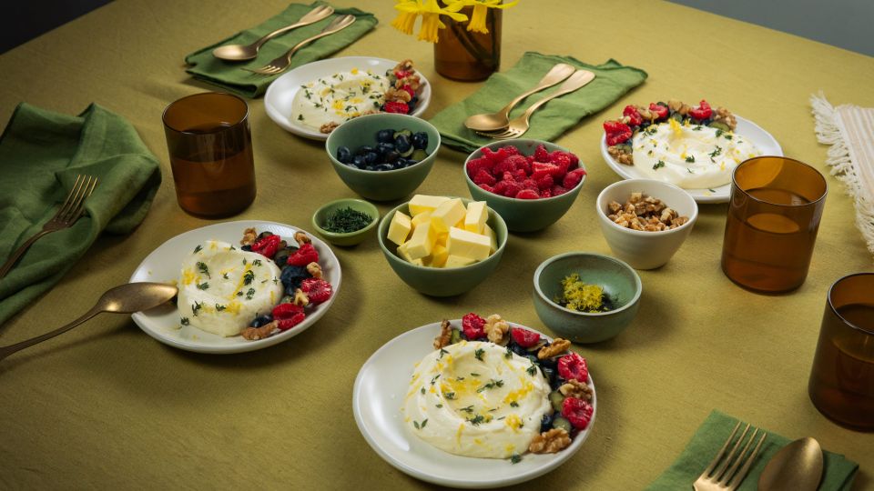 Auf einem gedeckten Tisch stehen mehrere Teller mit weißer Mousse, garniert mit Beeren und Nüssen. Daneben befinden sich kleine Schalen mit Butterstücken, Beeren und Nüssen sowie braune Gläser und grüne Stoffservietten.