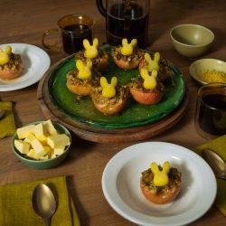 Mehrere halbierte, gebackene Äpfel mit knuspriger Füllung stehen auf Tellern und einer Servierplatte; darauf sitzen kleine Butterfiguren in Hasenform. Auf dem Holztisch stehen außerdem Schalen mit Butterwürfeln und Zitronenabrieb, Tassen mit dunklem Getränk sowie grüne Stoffservietten und Besteck.