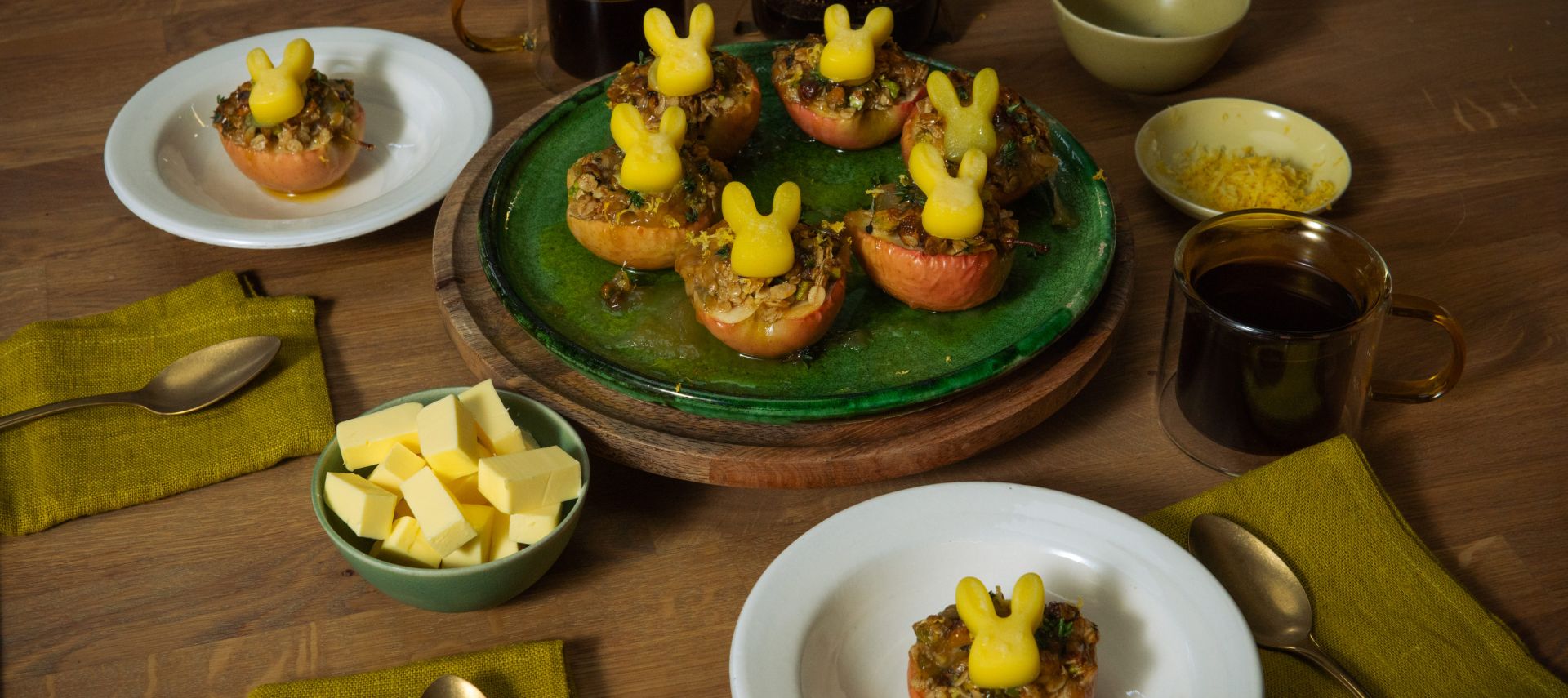 Mehrere halbierte, gebackene Äpfel mit knuspriger Füllung stehen auf Tellern und einer Servierplatte; darauf sitzen kleine Butterfiguren in Hasenform. Auf dem Holztisch stehen außerdem Schalen mit Butterwürfeln und Zitronenabrieb, Tassen mit dunklem Getränk sowie grüne Stoffservietten und Besteck.