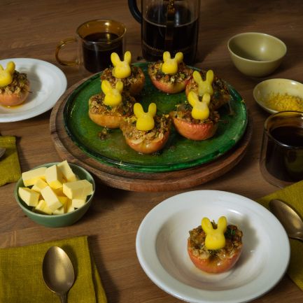 Mehrere halbierte, gebackene Äpfel mit knuspriger Füllung stehen auf Tellern und einer Servierplatte; darauf sitzen kleine Butterfiguren in Hasenform. Auf dem Holztisch stehen außerdem Schalen mit Butterwürfeln und Zitronenabrieb, Tassen mit dunklem Getränk sowie grüne Stoffservietten und Besteck.