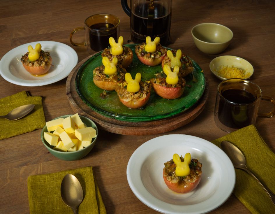 Mehrere halbierte, gebackene Äpfel mit knuspriger Füllung stehen auf Tellern und einer Servierplatte; darauf sitzen kleine Butterfiguren in Hasenform. Auf dem Holztisch stehen außerdem Schalen mit Butterwürfeln und Zitronenabrieb, Tassen mit dunklem Getränk sowie grüne Stoffservietten und Besteck.
