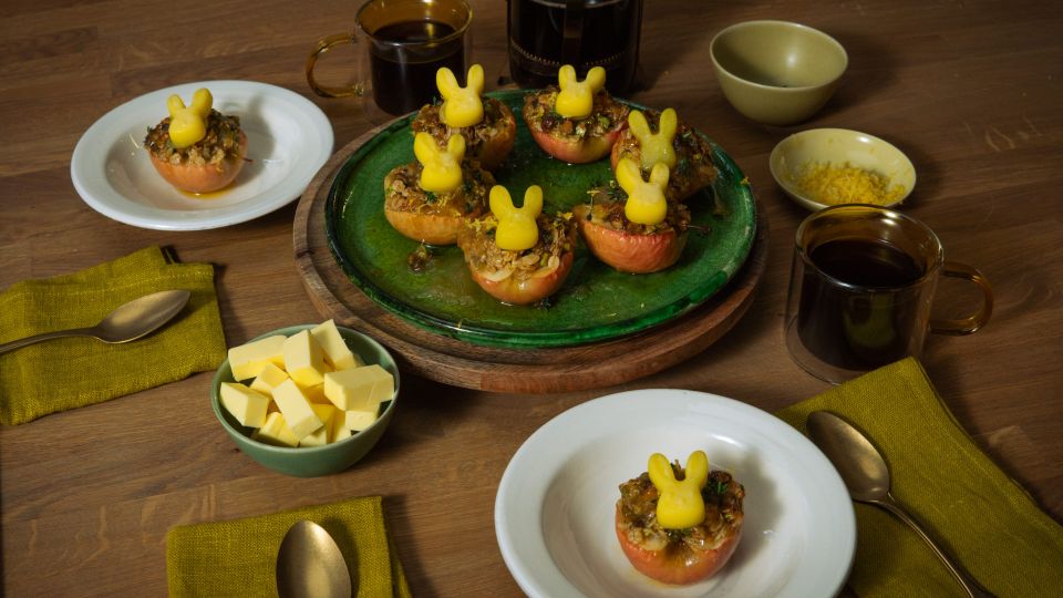 Mehrere halbierte, gebackene Äpfel mit knuspriger Füllung stehen auf Tellern und einer Servierplatte; darauf sitzen kleine Butterfiguren in Hasenform. Auf dem Holztisch stehen außerdem Schalen mit Butterwürfeln und Zitronenabrieb, Tassen mit dunklem Getränk sowie grüne Stoffservietten und Besteck.
