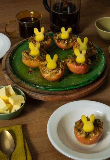 Mehrere halbierte, gebackene Äpfel mit knuspriger Füllung stehen auf Tellern und einer Servierplatte; darauf sitzen kleine Butterfiguren in Hasenform. Auf dem Holztisch stehen außerdem Schalen mit Butterwürfeln und Zitronenabrieb, Tassen mit dunklem Getränk sowie grüne Stoffservietten und Besteck.