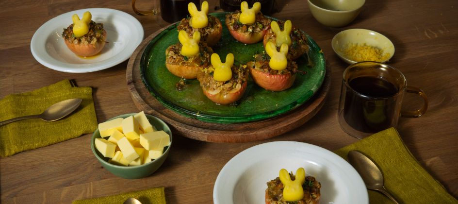 Mehrere halbierte, gebackene Äpfel mit knuspriger Füllung stehen auf Tellern und einer Servierplatte; darauf sitzen kleine Butterfiguren in Hasenform. Auf dem Holztisch stehen außerdem Schalen mit Butterwürfeln und Zitronenabrieb, Tassen mit dunklem Getränk sowie grüne Stoffservietten und Besteck.