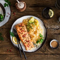 Ein Teller mit gebratenem Lachsfilet, serviert neben würzigem Bratreis und garniert mit Zitronenscheiben und frischen Kräutern, steht auf einem Holztisch. Daneben liegen kleine Schalen mit Zitronenstücken, Kräutern und Gewürzen sowie Besteck und ein Getränkeglas.