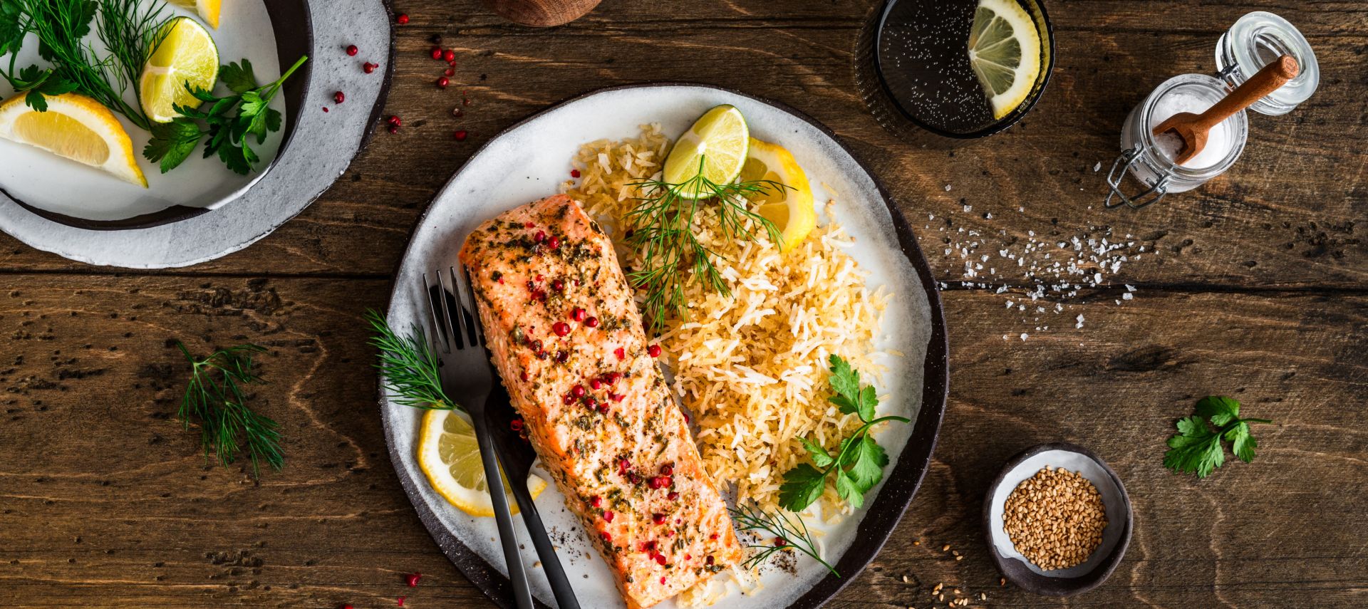 Ein Teller mit gebratenem Lachsfilet, serviert neben würzigem Bratreis und garniert mit Zitronenscheiben und frischen Kräutern, steht auf einem Holztisch. Daneben liegen kleine Schalen mit Zitronenstücken, Kräutern und Gewürzen sowie Besteck und ein Getränkeglas.