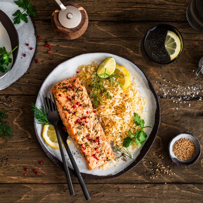 Ein Teller mit gebratenem Lachsfilet, serviert neben würzigem Bratreis und garniert mit Zitronenscheiben und frischen Kräutern, steht auf einem Holztisch. Daneben liegen kleine Schalen mit Zitronenstücken, Kräutern und Gewürzen sowie Besteck und ein Getränkeglas.