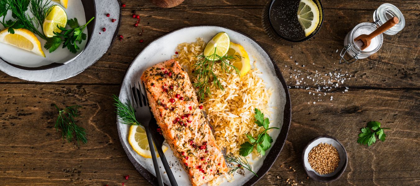 Ein Teller mit gebratenem Lachsfilet, serviert neben würzigem Bratreis und garniert mit Zitronenscheiben und frischen Kräutern, steht auf einem Holztisch. Daneben liegen kleine Schalen mit Zitronenstücken, Kräutern und Gewürzen sowie Besteck und ein Getränkeglas.