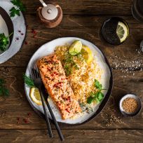 Ein Teller mit gebratenem Lachsfilet, serviert neben würzigem Bratreis und garniert mit Zitronenscheiben und frischen Kräutern, steht auf einem Holztisch. Daneben liegen kleine Schalen mit Zitronenstücken, Kräutern und Gewürzen sowie Besteck und ein Getränkeglas.