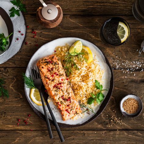 Ein Teller mit gebratenem Lachsfilet, serviert neben würzigem Bratreis und garniert mit Zitronenscheiben und frischen Kräutern, steht auf einem Holztisch. Daneben liegen kleine Schalen mit Zitronenstücken, Kräutern und Gewürzen sowie Besteck und ein Getränkeglas.