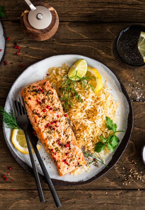Ein Teller mit gebratenem Lachsfilet, serviert neben würzigem Bratreis und garniert mit Zitronenscheiben und frischen Kräutern, steht auf einem Holztisch. Daneben liegen kleine Schalen mit Zitronenstücken, Kräutern und Gewürzen sowie Besteck und ein Getränkeglas.