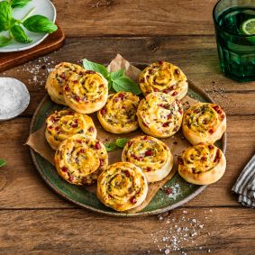 Auf einem rustikalen Holztisch steht ein Teller voller herzhafter Flammkuchenschnecken, garniert mit Kräutern. Daneben liegen eine Schale mit grobem Salz, frische Basilikumblätter, ein grünes Trinkglas mit Limettenscheibe und ein gestreiftes Tuch.