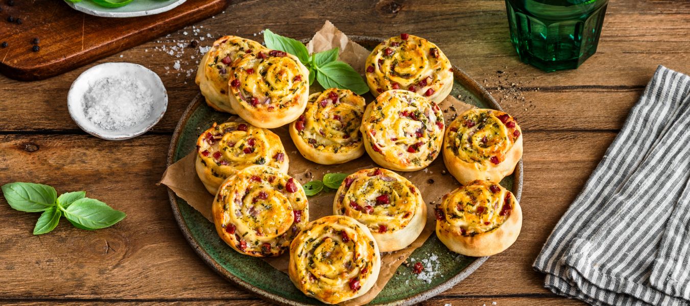 Auf einem rustikalen Holztisch steht ein Teller voller herzhafter Flammkuchenschnecken, garniert mit Kräutern. Daneben liegen eine Schale mit grobem Salz, frische Basilikumblätter, ein grünes Trinkglas mit Limettenscheibe und ein gestreiftes Tuch.