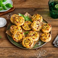 Auf einem rustikalen Holztisch steht ein Teller voller herzhafter Flammkuchenschnecken, garniert mit Kräutern. Daneben liegen eine Schale mit grobem Salz, frische Basilikumblätter, ein grünes Trinkglas mit Limettenscheibe und ein gestreiftes Tuch.