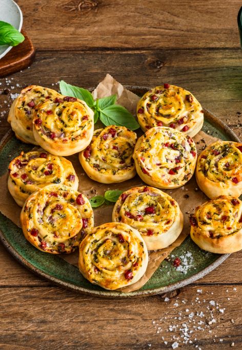 Auf einem rustikalen Holztisch steht ein Teller voller herzhafter Flammkuchenschnecken, garniert mit Kräutern. Daneben liegen eine Schale mit grobem Salz, frische Basilikumblätter, ein grünes Trinkglas mit Limettenscheibe und ein gestreiftes Tuch.