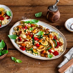Ein großer Teller mit bunt gemischtem Grillgemüse und Perlcouscous steht auf einem Holztisch. Das Gericht ist mit frischem Basilikum garniert. Daneben stehen ein kleinerer Teller mit einer weiteren Portion, ein Teller mit Zitronenscheiben, eine Salzschale und Küchenutensilien.