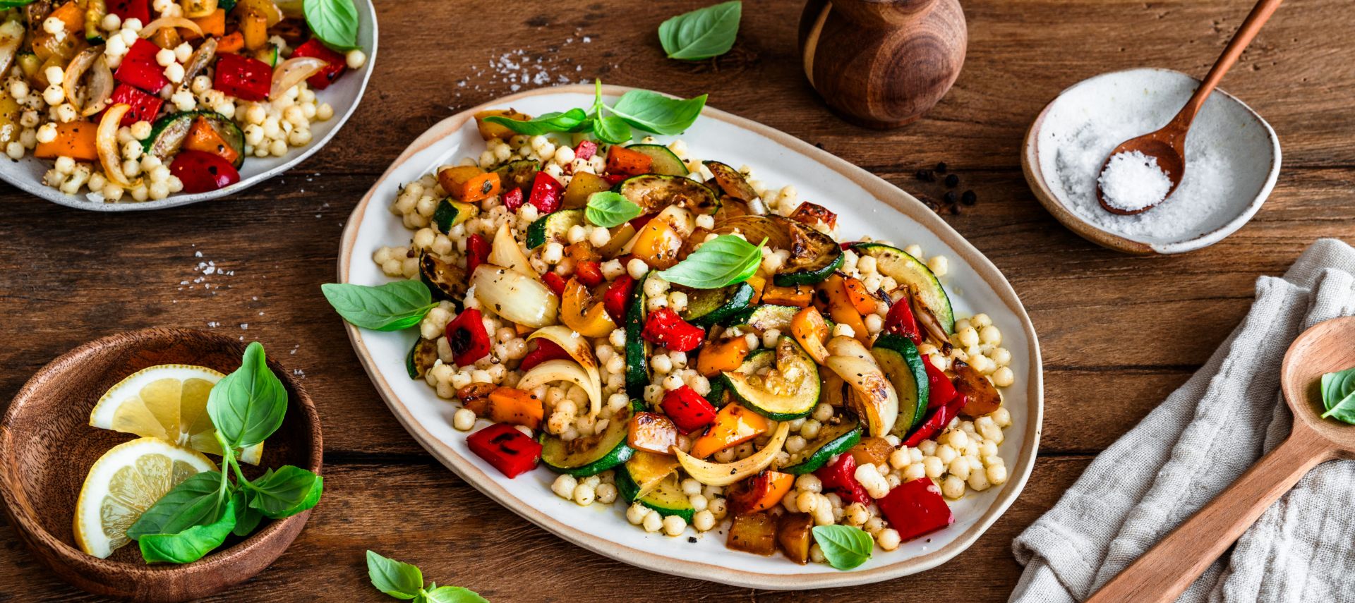 Ein großer Teller mit bunt gemischtem Grillgemüse und Perlcouscous steht auf einem Holztisch. Das Gericht ist mit frischem Basilikum garniert. Daneben stehen ein kleinerer Teller mit einer weiteren Portion, ein Teller mit Zitronenscheiben, eine Salzschale und Küchenutensilien.