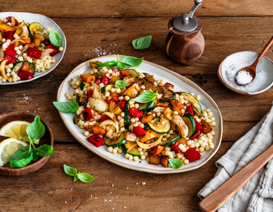 Ein großer Teller mit bunt gemischtem Grillgemüse und Perlcouscous steht auf einem Holztisch. Das Gericht ist mit frischem Basilikum garniert. Daneben stehen ein kleinerer Teller mit einer weiteren Portion, ein Teller mit Zitronenscheiben, eine Salzschale und Küchenutensilien.