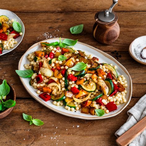 Ein großer Teller mit bunt gemischtem Grillgemüse und Perlcouscous steht auf einem Holztisch. Das Gericht ist mit frischem Basilikum garniert. Daneben stehen ein kleinerer Teller mit einer weiteren Portion, ein Teller mit Zitronenscheiben, eine Salzschale und Küchenutensilien.
