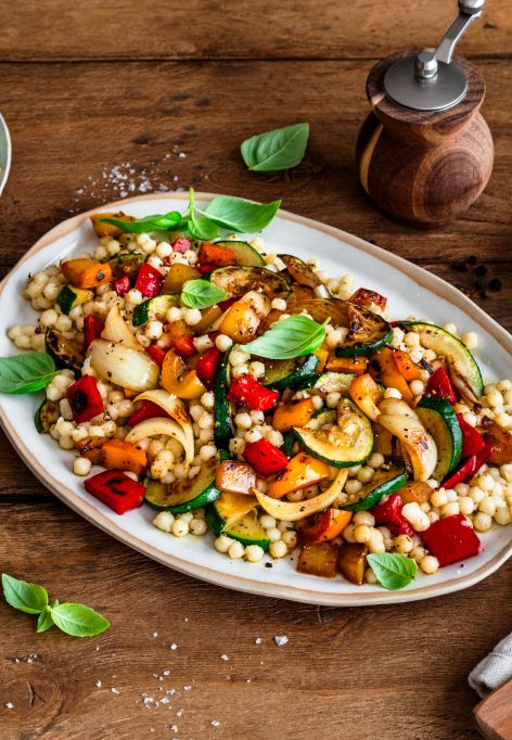 Ein großer Teller mit bunt gemischtem Grillgemüse und Perlcouscous steht auf einem Holztisch. Das Gericht ist mit frischem Basilikum garniert. Daneben stehen ein kleinerer Teller mit einer weiteren Portion, ein Teller mit Zitronenscheiben, eine Salzschale und Küchenutensilien.