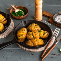 In zwei kleinen gusseisernen Pfannen liegen goldbraun gebackene Hasselback-Kartoffeln, bestreut mit frischen Kräutern. Die Pfannen stehen auf Untersetzern auf einem Holztisch. Daneben befinden sich Schalen mit Schnittlauch und grobem Salz, ein Pfeffermühle, Besteck und einzelne Basilikumblätter.