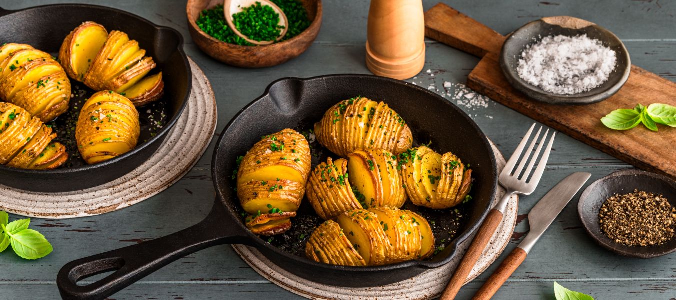 In zwei kleinen gusseisernen Pfannen liegen goldbraun gebackene Hasselback-Kartoffeln, bestreut mit frischen Kräutern. Die Pfannen stehen auf Untersetzern auf einem Holztisch. Daneben befinden sich Schalen mit Schnittlauch und grobem Salz, ein Pfeffermühle, Besteck und einzelne Basilikumblätter.