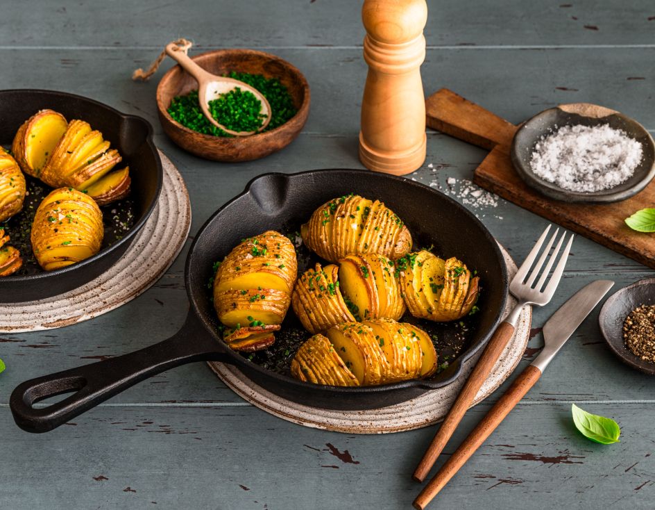 In zwei kleinen gusseisernen Pfannen liegen goldbraun gebackene Hasselback-Kartoffeln, bestreut mit frischen Kräutern. Die Pfannen stehen auf Untersetzern auf einem Holztisch. Daneben befinden sich Schalen mit Schnittlauch und grobem Salz, ein Pfeffermühle, Besteck und einzelne Basilikumblätter.