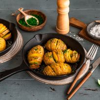In zwei kleinen gusseisernen Pfannen liegen goldbraun gebackene Hasselback-Kartoffeln, bestreut mit frischen Kräutern. Die Pfannen stehen auf Untersetzern auf einem Holztisch. Daneben befinden sich Schalen mit Schnittlauch und grobem Salz, ein Pfeffermühle, Besteck und einzelne Basilikumblätter.