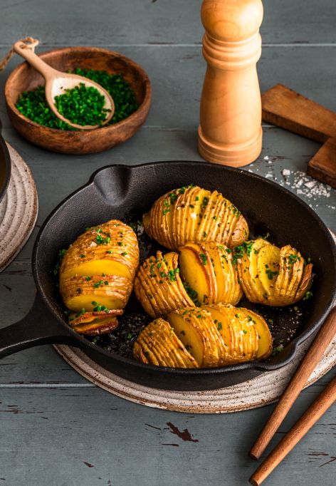 In zwei kleinen gusseisernen Pfannen liegen goldbraun gebackene Hasselback-Kartoffeln, bestreut mit frischen Kräutern. Die Pfannen stehen auf Untersetzern auf einem Holztisch. Daneben befinden sich Schalen mit Schnittlauch und grobem Salz, ein Pfeffermühle, Besteck und einzelne Basilikumblätter.