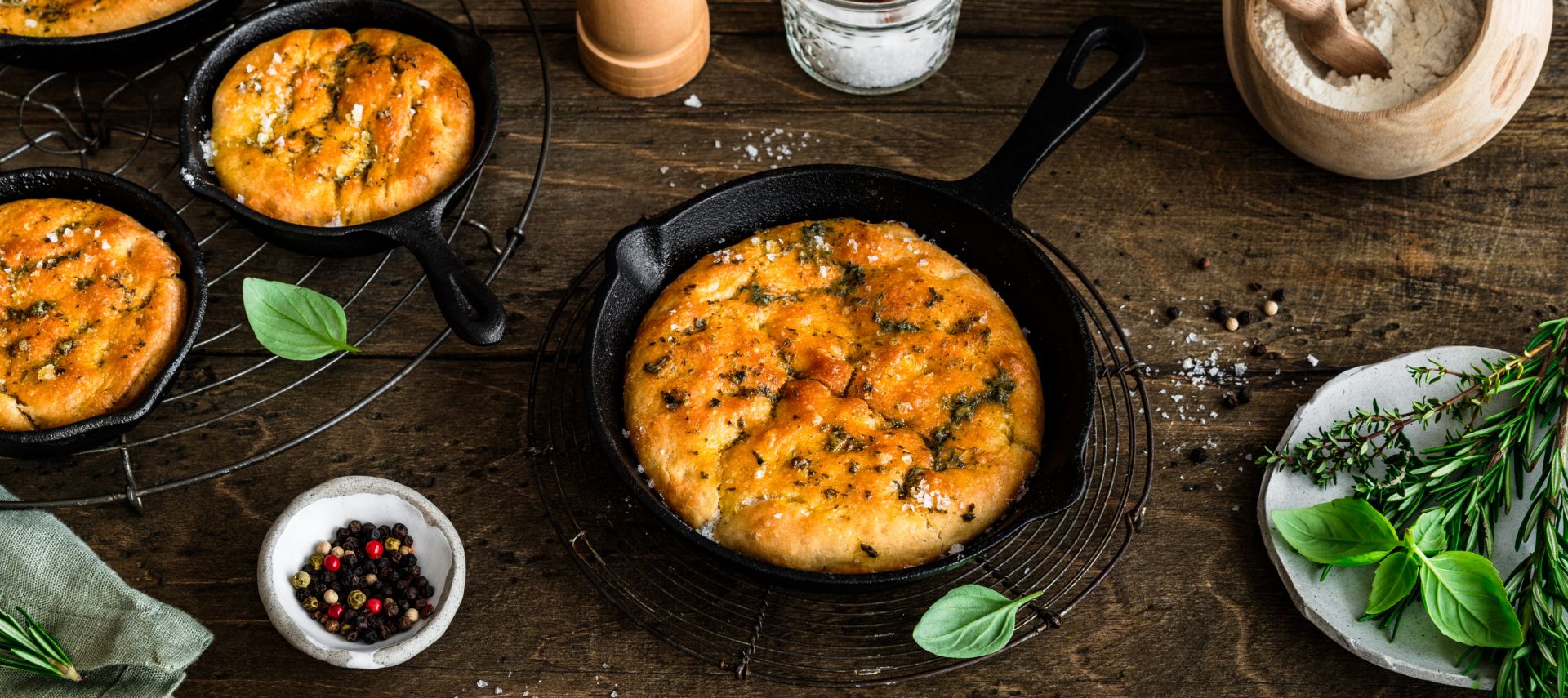 In kleinen gusseisernen Pfannen liegt frisch gebackene Focaccia, goldbraun und mit Kräutern bestreut. Die Pfannen stehen auf einem rustikalen Holztisch. Daneben befinden sich Schalen mit Mehl und Salz, eine Pfeffermühle, frische Kräuterzweige sowie ein Küchentuch.