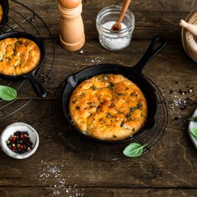 In kleinen gusseisernen Pfannen liegt frisch gebackene Focaccia, goldbraun und mit Kräutern bestreut. Die Pfannen stehen auf einem rustikalen Holztisch. Daneben befinden sich Schalen mit Mehl und Salz, eine Pfeffermühle, frische Kräuterzweige sowie ein Küchentuch.