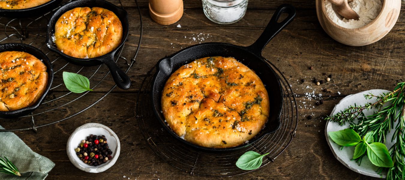 In kleinen gusseisernen Pfannen liegt frisch gebackene Focaccia, goldbraun und mit Kräutern bestreut. Die Pfannen stehen auf einem rustikalen Holztisch. Daneben befinden sich Schalen mit Mehl und Salz, eine Pfeffermühle, frische Kräuterzweige sowie ein Küchentuch.