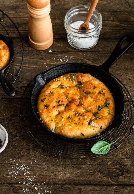 In kleinen gusseisernen Pfannen liegt frisch gebackene Focaccia, goldbraun und mit Kräutern bestreut. Die Pfannen stehen auf einem rustikalen Holztisch. Daneben befinden sich Schalen mit Mehl und Salz, eine Pfeffermühle, frische Kräuterzweige sowie ein Küchentuch.