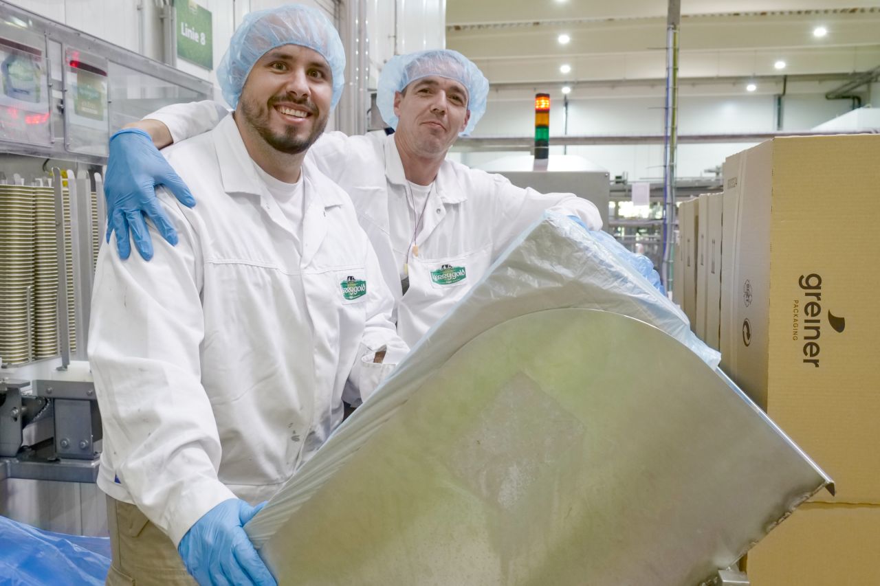 Zwei Personen in weißen Kitteln stehen in einer Produktionshalle und lachen in die Kamera.
